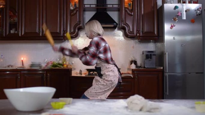 快乐的高加索年轻女子在家中厨房跳舞的镜头。围裙里开朗美丽的女士在室内做饭的肖像。生活方式和幸福。