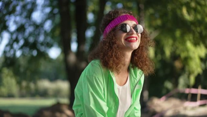 中拍肖像快乐的女人戴上墨镜笑着看着相机。快乐放松的高加索复古女士在夏日公园慢动作摆姿势。生活方式和幸