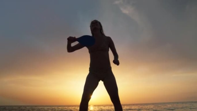 日落时在海滩上享受桨网球的女人的剪影。独自练习