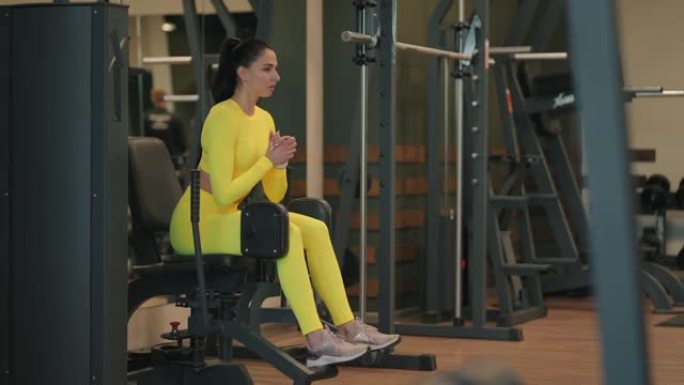 强壮的西班牙裔女运动员在模拟器上做腿部稀释练习。穿着黄色运动服的运动健身房的黑发女人在腿部压榨机上锻