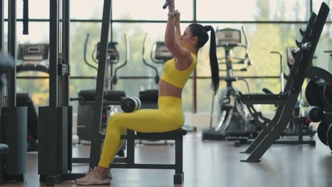 一个穿着黄色西装的年轻黑发女人正在模拟器上锻炼背部的肌肉。西班牙裔妇女在模拟器上拉绳子。模拟引体向上