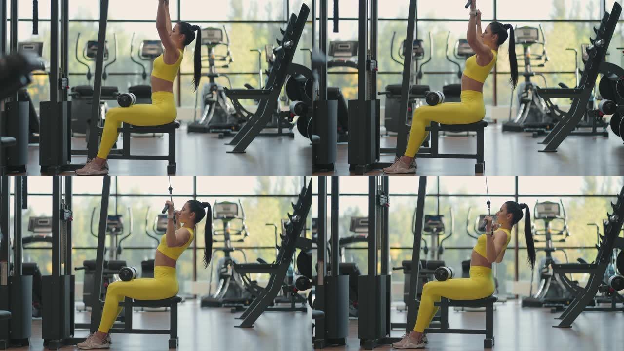 一个穿着黄色西装的年轻黑发女人正在模拟器上锻炼背部的肌肉。西班牙裔妇女在模拟器上拉绳子。模拟引体向上