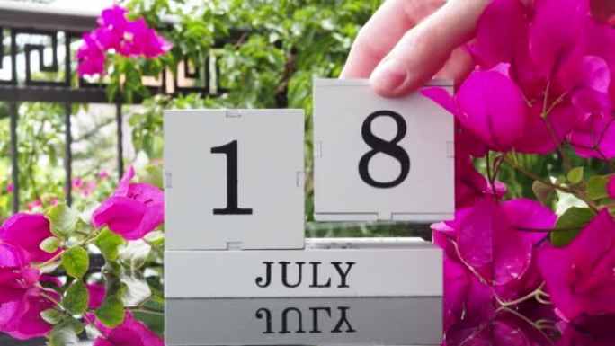 一个木制日历，有一个重要的活动，7月18日在一张有芙蓉花的桌子上，女人的手在日历上设定了日期。将日历