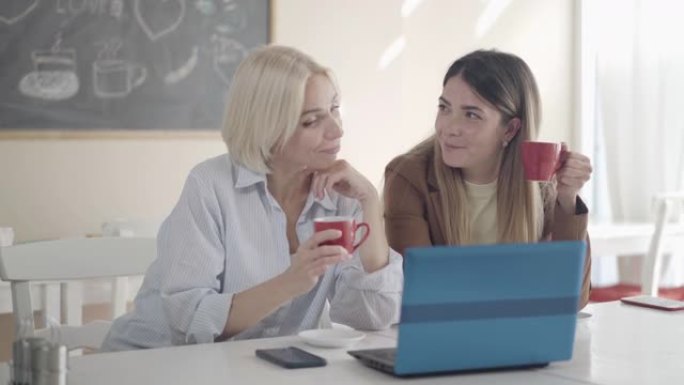 自信的年轻白人妇女休息时在午餐室闲聊。两个漂亮的同事坐在笔记本电脑的午餐室里聊天和喝茶或咖啡的肖像。