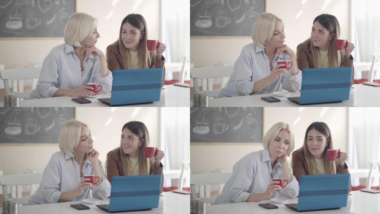 自信的年轻白人妇女休息时在午餐室闲聊。两个漂亮的同事坐在笔记本电脑的午餐室里聊天和喝茶或咖啡的肖像。