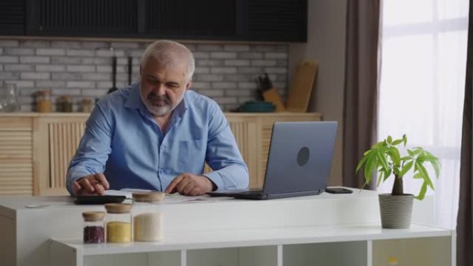 老人正在计算自己的财务状况，坐在家里的厨房里，用计算器计数并在书本上做笔记，使用笔记本电脑