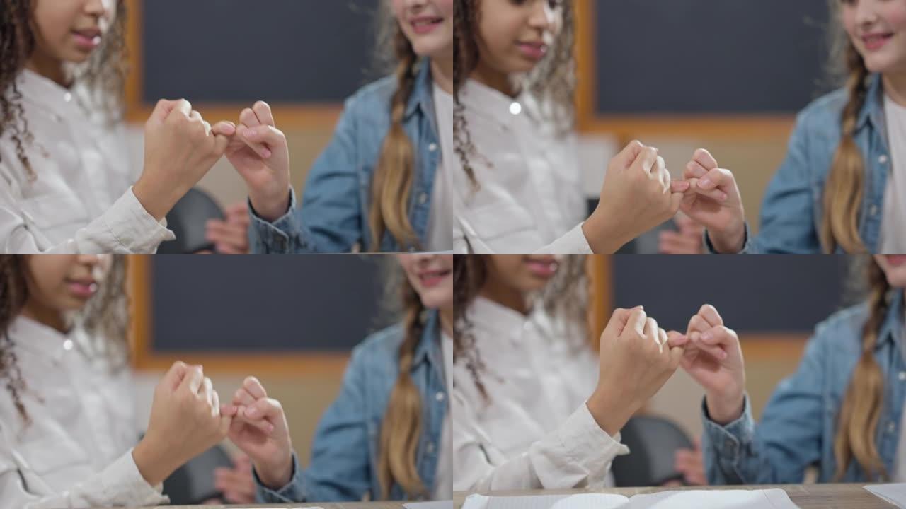 教室里无法辨认的高加索和非裔美国女学生的特写小指誓言。快乐的朋友女孩坐在教室的桌子上摇晃小手指。