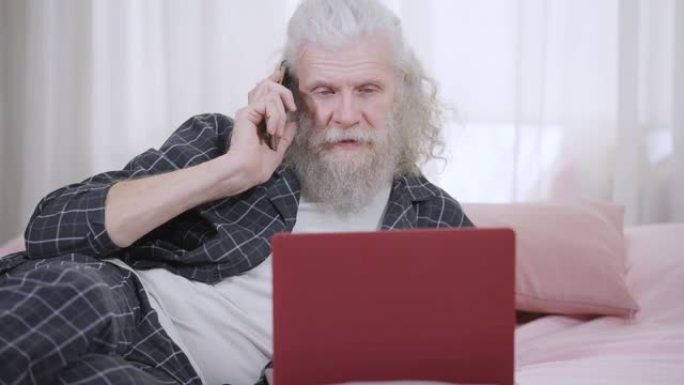长胡子的英俊高级白发男子的肖像躺在笔记本电脑的床上在电话上聊天。穿着睡衣的轻松自信的高加索退休人员在