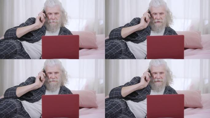 长胡子的英俊高级白发男子的肖像躺在笔记本电脑的床上在电话上聊天。穿着睡衣的轻松自信的高加索退休人员在