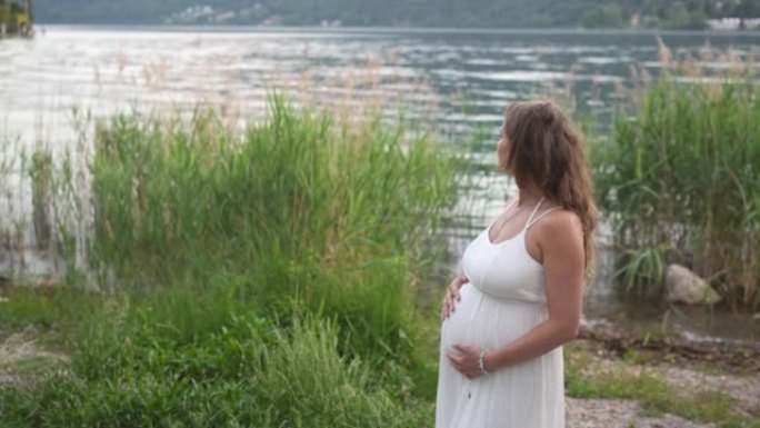 穿着白色连衣裙的孕妇抱着肚子在湖边放松，可以看到山景