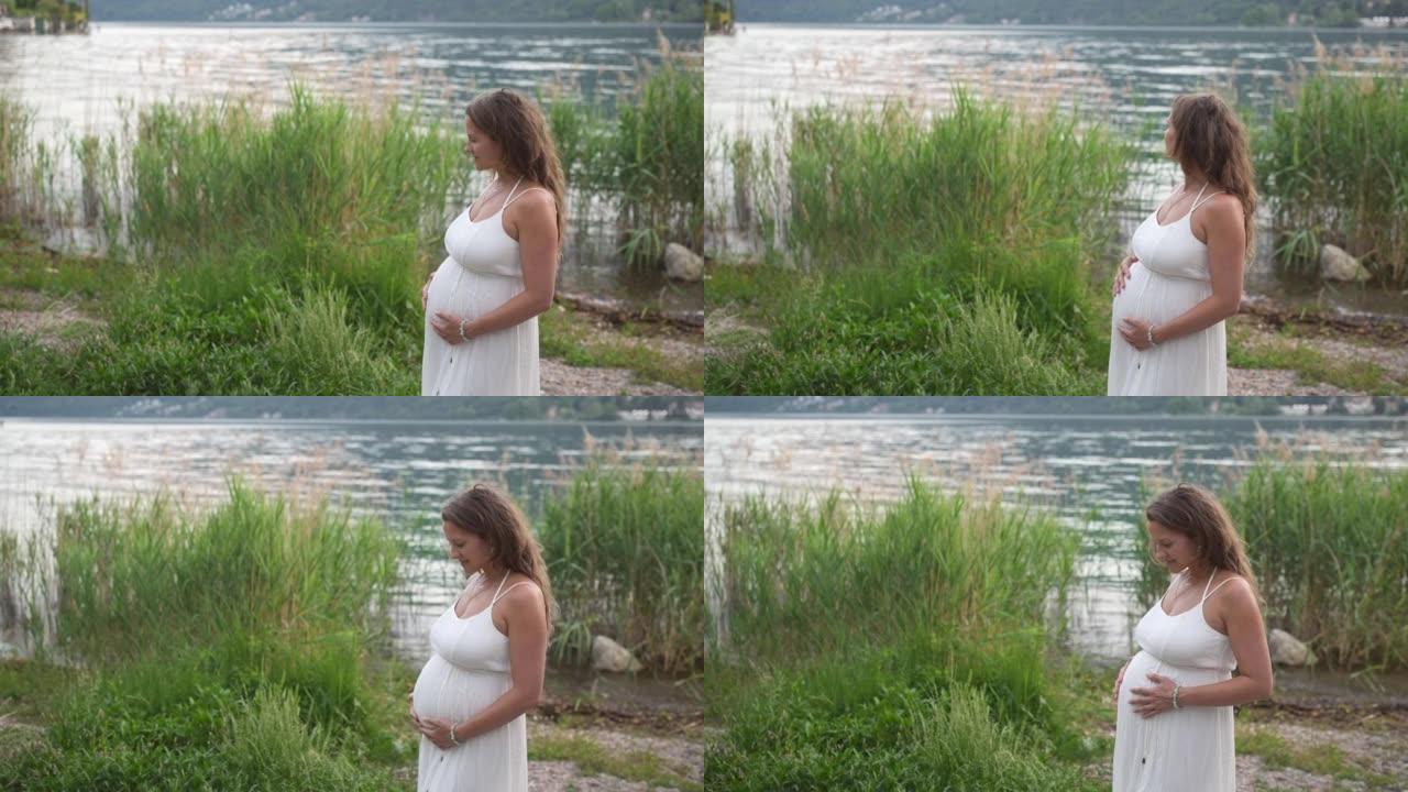 穿着白色连衣裙的孕妇抱着肚子在湖边放松，可以看到山景