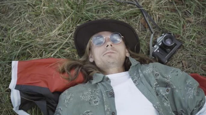 戴着墨镜的放松嬉皮士男子的特写镜头，躺在绿草丛中，对着镜头微笑。粗心的高加索20世纪60年代家伙在夏