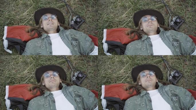戴着墨镜的放松嬉皮士男子的特写镜头，躺在绿草丛中，对着镜头微笑。粗心的高加索20世纪60年代家伙在夏