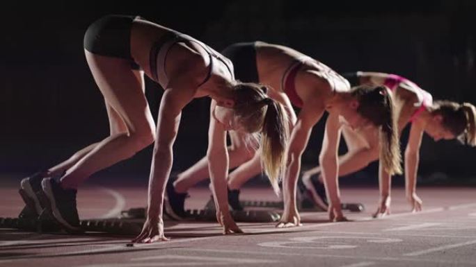 一排跑步者女子在开始比赛前蹲在起跑位置。雌性从体育场的起跑鞋开始，在黑暗中，聚光灯在慢动作中。