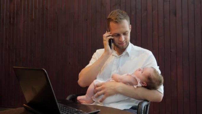 年轻的父亲男子试图在笔记本电脑上工作，并与手机客户端交谈，同时手握着熟睡的女儿孩子
