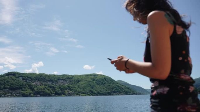 女人坐在木制栏杆上，使用智能手机俯瞰湖泊和绿色山脉