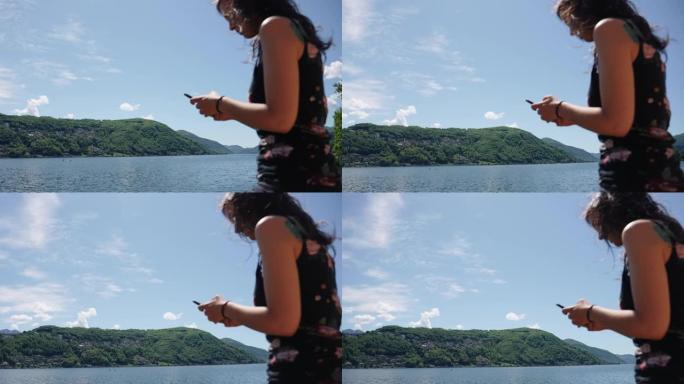 女人坐在木制栏杆上，使用智能手机俯瞰湖泊和绿色山脉