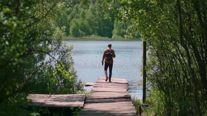 一个背着背包的年轻人沿着森林中湖岸的码头散步，用手机拍摄散步和风景如画的自然景色。游客欣赏美丽的风景