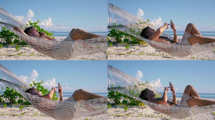 男子用电话躺在海滩上两棵棕榈树之间的吊床上