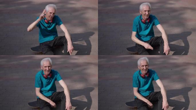 高角度视角积极的高级运动员坐在阳光公园小巷的滑板上，慢动作向摄像机致敬。快乐英俊的高加索退休人员在户