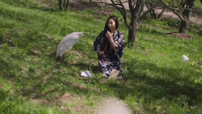 亚洲苗条的年轻美女坐在阳光明媚的公园绿色的春天草地上抚摸头发。自信优雅的日本千禧一代在户外阳光下休息