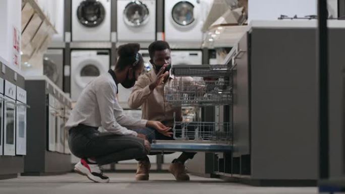 带有口罩的美国黑人已婚夫妇正在家用电器商店中选择洗碗机，检查厨房设备