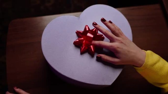 女孩正在为情人节，生日或周年纪念日的亲人准备惊喜。将红色蝴蝶结贴在心形礼品盒上的手的特写镜头