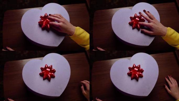女孩正在为情人节，生日或周年纪念日的亲人准备惊喜。将红色蝴蝶结贴在心形礼品盒上的手的特写镜头