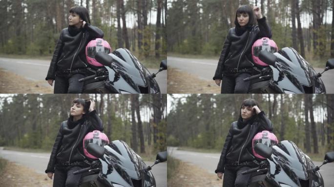 美丽的高加索女性摩托车手站在黑色摩托车上，戴着粉色头盔，目光移开。自信的年轻黑发女人在乡下路边。