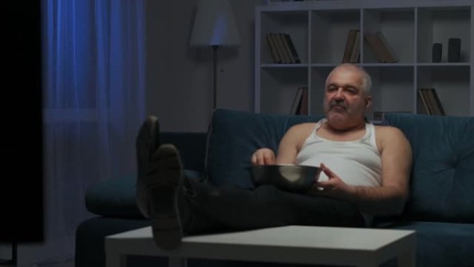 一个留着小胡子的男人正在电视上看动作片，吃爆米花。屏幕上明亮的闪光。