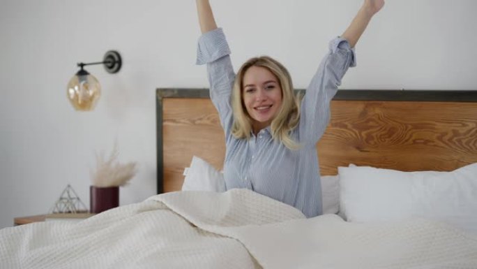 快乐的高加索女人早上在家躺在床上伸展。快乐微笑的美丽女士在卧室里醒来看着相机的肖像。休闲和美容概念。