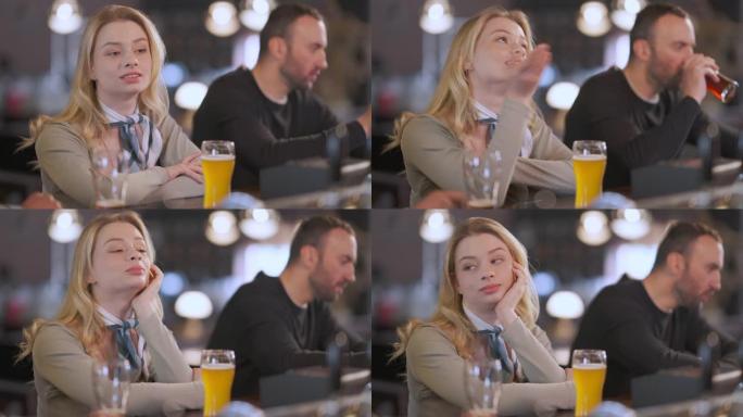 年轻漂亮的女人与坐在酒吧柜台的男人聊天的肖像，背景模糊的客户在喝啤酒。放松的高加索女友在室内酒吧和男