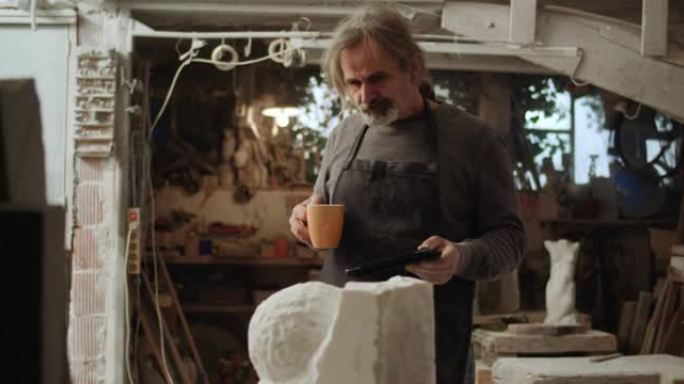 高级男雕塑家在他的工作室里用平板电脑喝了一口茶