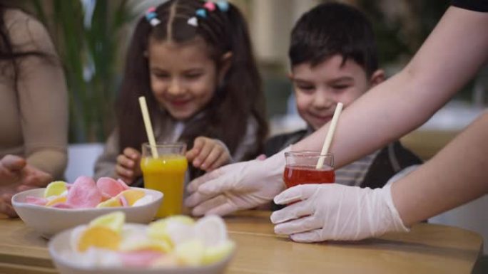 女服务员双手为坐在海鲜餐厅餐桌上的快乐中东男孩和女孩提供黄色和红色的果汁。积极笑兄妹享受家庭休闲。慢