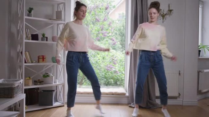 宽镜头肖像两个迷人美丽的同卵双胞胎在客厅看着相机跳舞。开朗自信的高加索姐妹周末在家里摆姿势很开心。