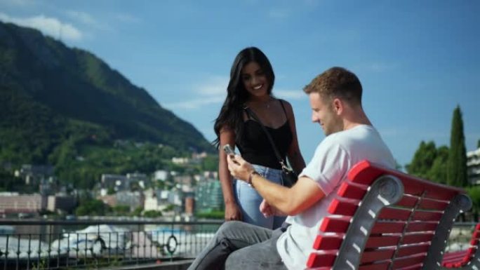 男人和女人在使用智能手机时在红色长凳上放松，可以看到湖边的笑声
