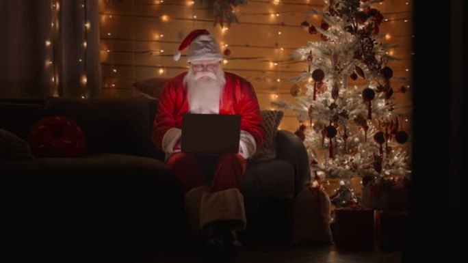 总体平面图前视图一个真正的圣诞老人在晚上用笔记本电脑工作，在圣诞树背景上的圣诞灯的灯光下戴着眼镜。远