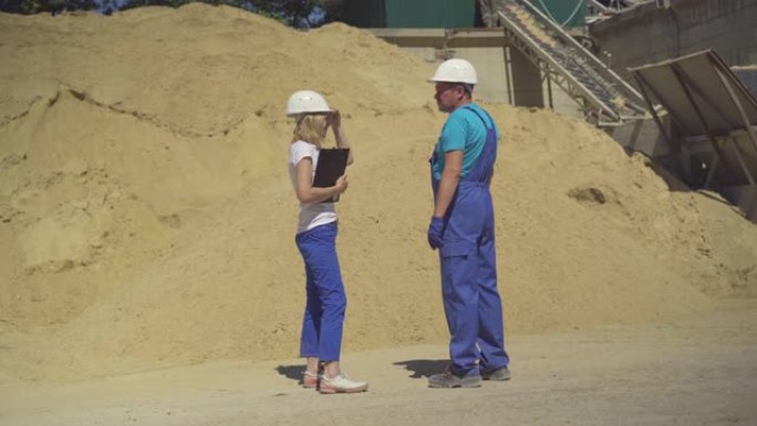 专业的女检查员和男工人站在巨大的沙堆前的制造现场交谈。水泥厂戴着安全帽的高加索男人和女人。