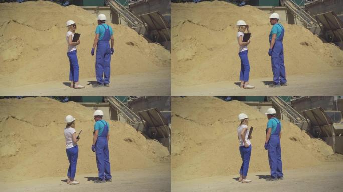 专业的女检查员和男工人站在巨大的沙堆前的制造现场交谈。水泥厂戴着安全帽的高加索男人和女人。