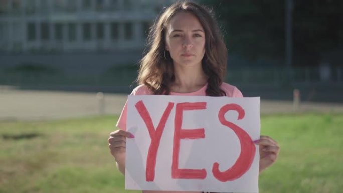 一个年轻的黑发女人在户外拿着Yes的海报。严肃的白人女性活动家举着标语站在阳光下。漂亮的抗议者在一个