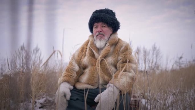 自信的土著老人的肖像坐在户外，坐在黄草的冬季草地上。高个子英俊的大胡子蒙古人在寒冷的日子里看着相机。