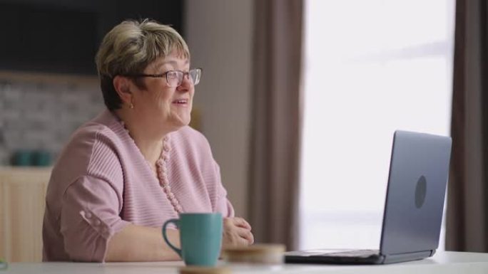 开朗的老太太通过笔记本电脑上的视频聊天与家人交流，老太太周末坐在家里的厨房里