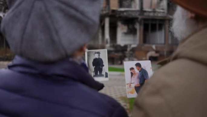 拍摄越过肩膀模糊的高级男子和妇女说话，看着旧照片站在废墟建筑的背景。乌克兰城市被炸毁的房子里绝望的夫