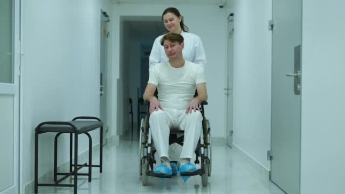宽镜头肖像积极的医生说话的病人推着轮椅在医院的走廊。前视图白种人护士支持瘫痪病人滚动移动辅助装置在诊