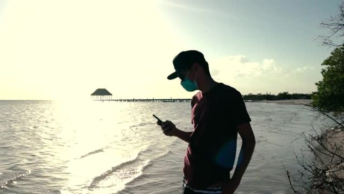 年轻人在日落时戴着口罩在海滩上使用电话