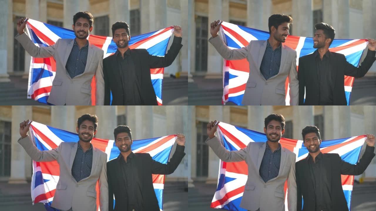两个穿着西装的快乐中东年轻人看着相机微笑着站在户外的英国国旗的中等镜头。城市中自信的帅哥在阳光下摆姿