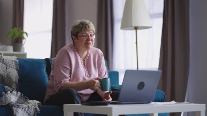 戴眼镜的快乐老太太通过与朋友或家人的在线视频聊天进行交流，女性退休人员坐在家里的沙发上
