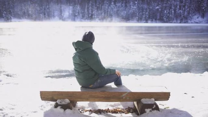 年轻人坐在白雪皑皑的湖边附近的木凳上欣赏美景