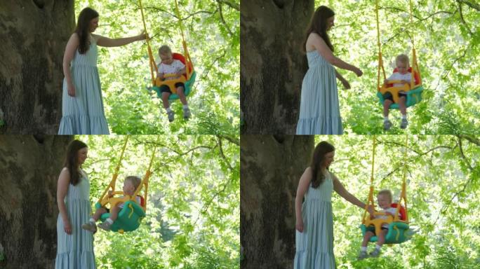 母亲带着快乐的小孩，在阳光明媚的夏日，在挂在树上的户外婴儿秋千上玩得很开心。儿童健康暑期活动。