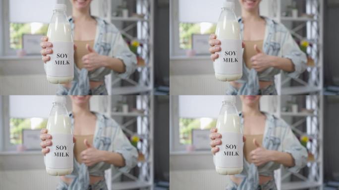 特写豆奶瓶，模糊的无法辨认的白人妇女在背景下示意拇指向上微笑。年轻苗条快乐的女士在家里摆姿势，室内喝